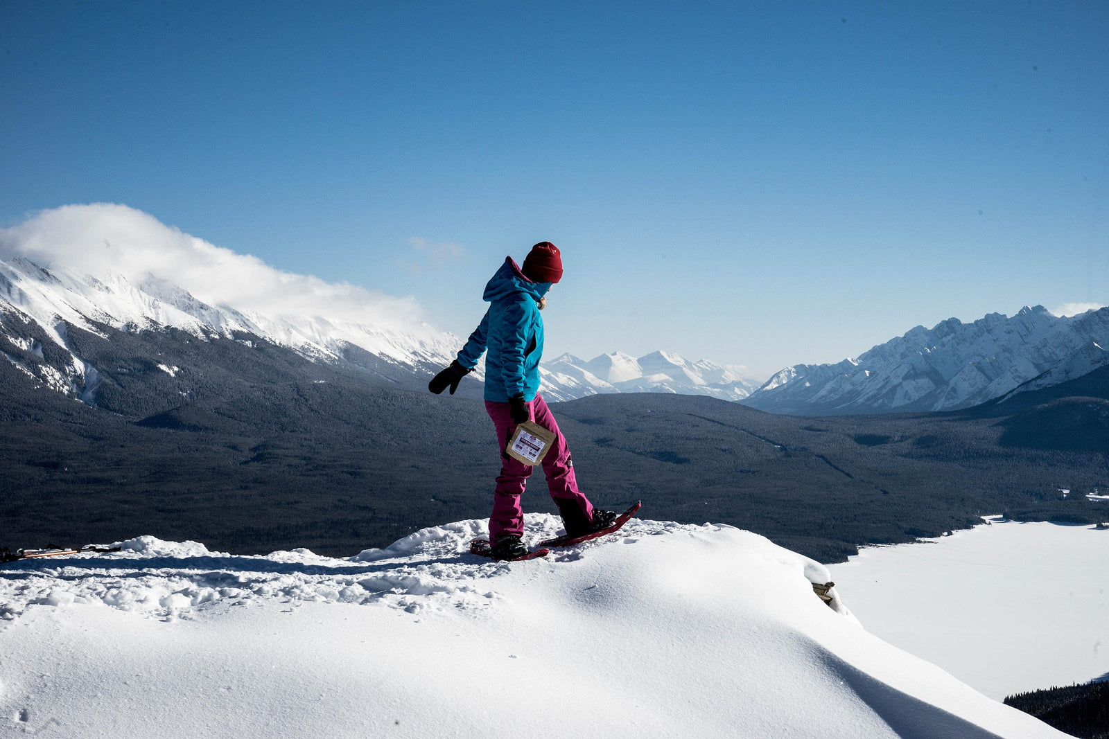 Woman holding Nomad Nutrition Ukrainian Borscht meal while snowshoeing across snowy winter trails with snow capped mountains in the background. Easy and instant dehydrated meal for winter activities.
