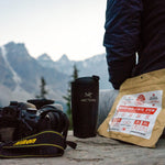 Nomad Nutrition Indian Red Lentil Stew 112g or 4oz meal size next to person, thermos, and camera. Scene overlooking blue lake and snow capped mountains. Convenient size for travelling.