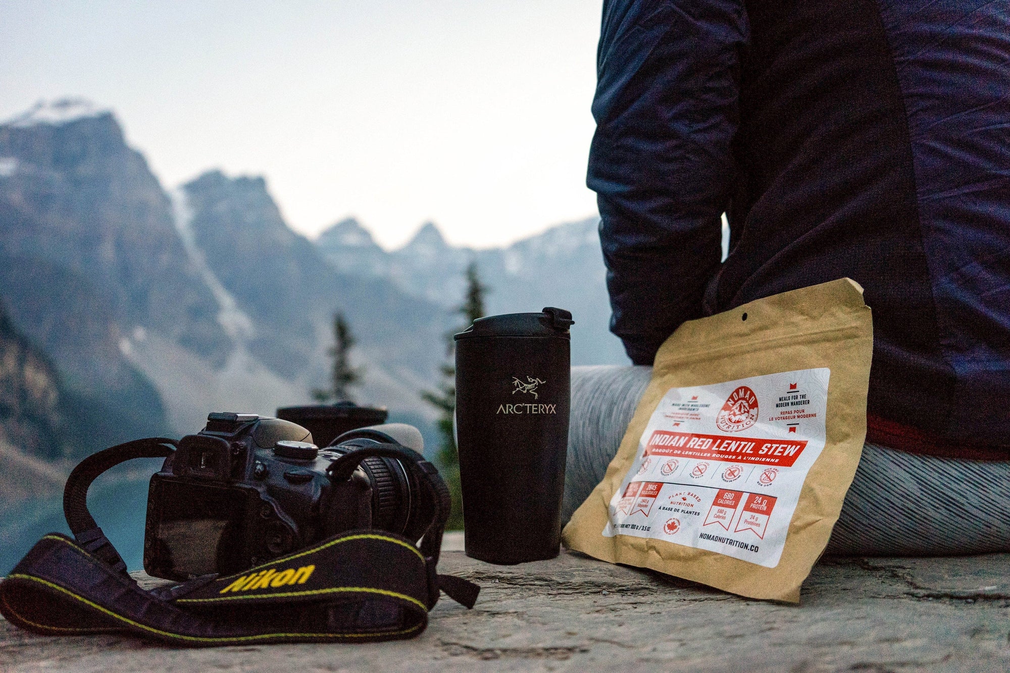 Nomad Nutrition Indian Red Lentil Stew 112g or 4oz meal size next to person, thermos, and camera. Scene overlooking blue lake and snow capped mountains. Convenient size for travelling.