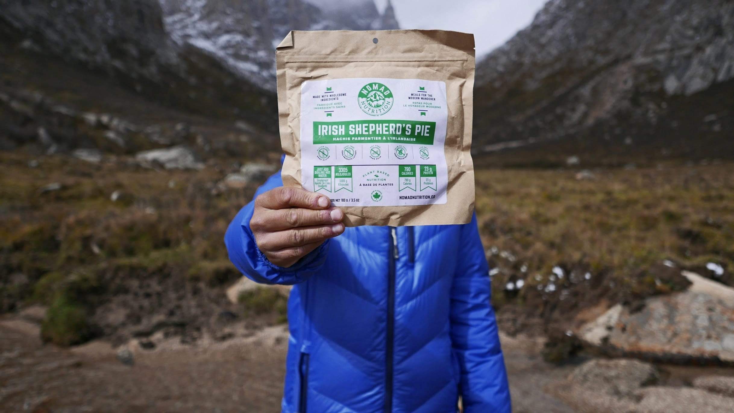 Person holding Irish Shepherd's Pie 112g/ 4oz, Person holding Irish Shepherd's Pie (112g/ 4oz), adehydrated vegan adventure plant-based, gluten-free meal outdoors surrounded by mountains. 