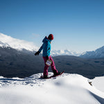Woman holding Nomad Nutrition Ukrainian Borscht meal while snowshoeing across snowy winter trails with snow capped mountains in the background. Easy and instant dehydrated meal for winter activities. 